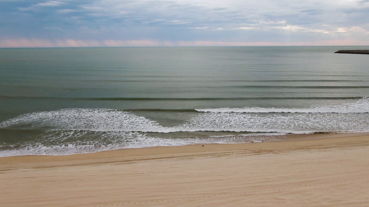 Aerial frame of the San Felipe shoreline with thin parallel wave lines offshore, a white breaking wave near the right edge, tan sand below, and no visible people.