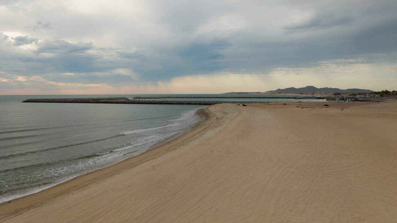 Opening aerial frame over San Felipe, BC, Mexico with empty sand in the foreground, calm water to the left, a long pier across the midground, distant mountains, heavy clouds, and no visible people.