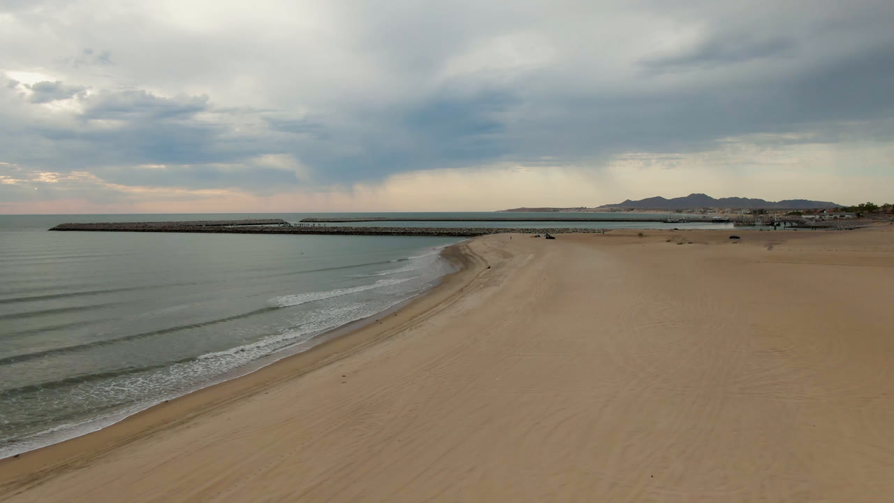 At one second, the San Felipe beach remains empty, with low waves lining the left shoreline, the pier stretching across the center distance, muted sand tones, cloudy light, and no visible people.