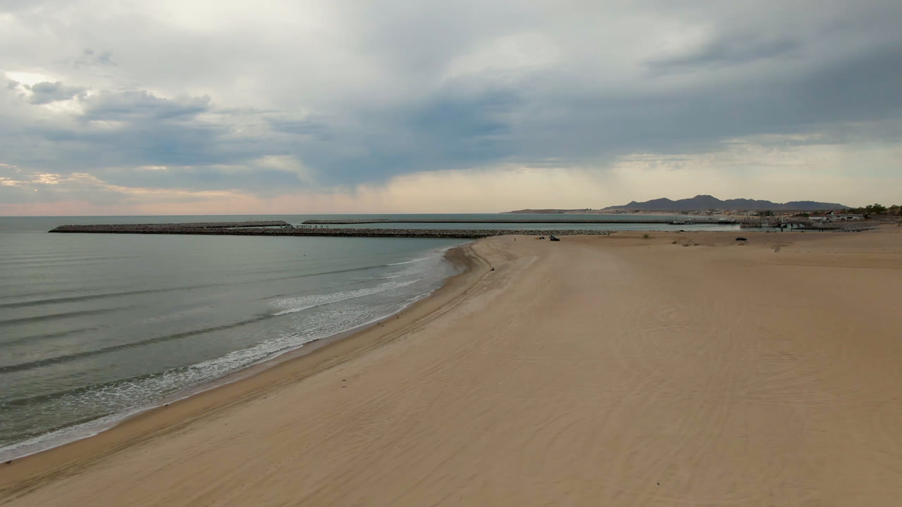 Empty beach beside calm water with a pier, mountains, and thick clouds in the distance.