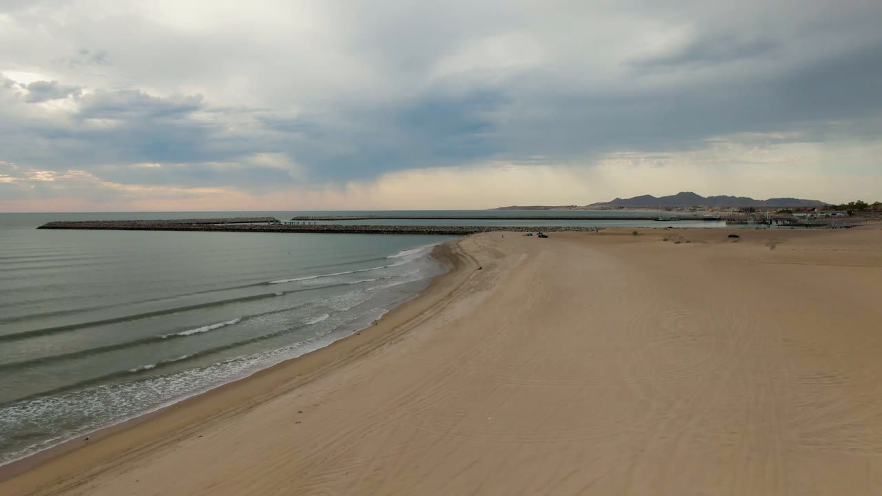 Opening aerial frame over San Felipe BC Mexico with muted water at left, a broad empty beach at right, a long pier across the midground, distant hills, heavy clouds, and no visible people.