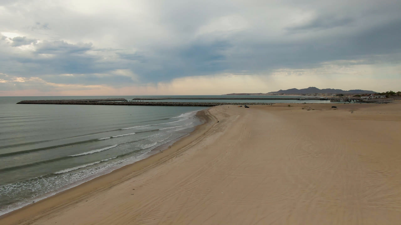 At one second, the San Felipe BC Mexico shoreline remains wide and empty, with small parallel waves nearer the lower left, the pier stretching across the bay, distant mountains, and no visible people.