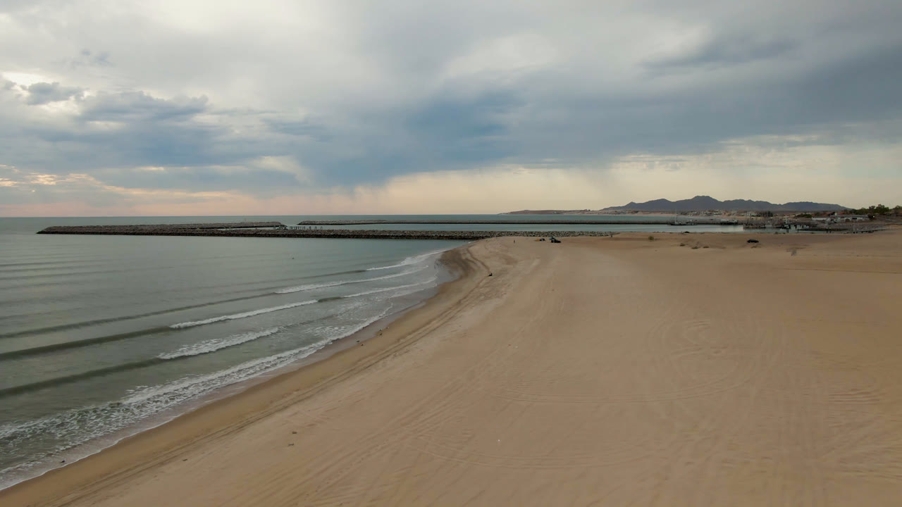 Curving beach beside calm water with pier, clouds, and low mountains beyond.