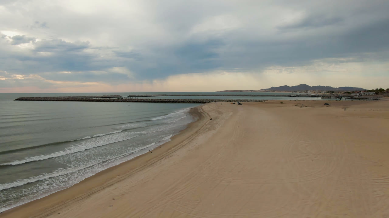 Opening aerial frame over San Felipe, BC, Mexico, with muted surf at left, a broad empty sand beach at right, a long pier on the horizon, distant hills, gray clouds, and no visible people.