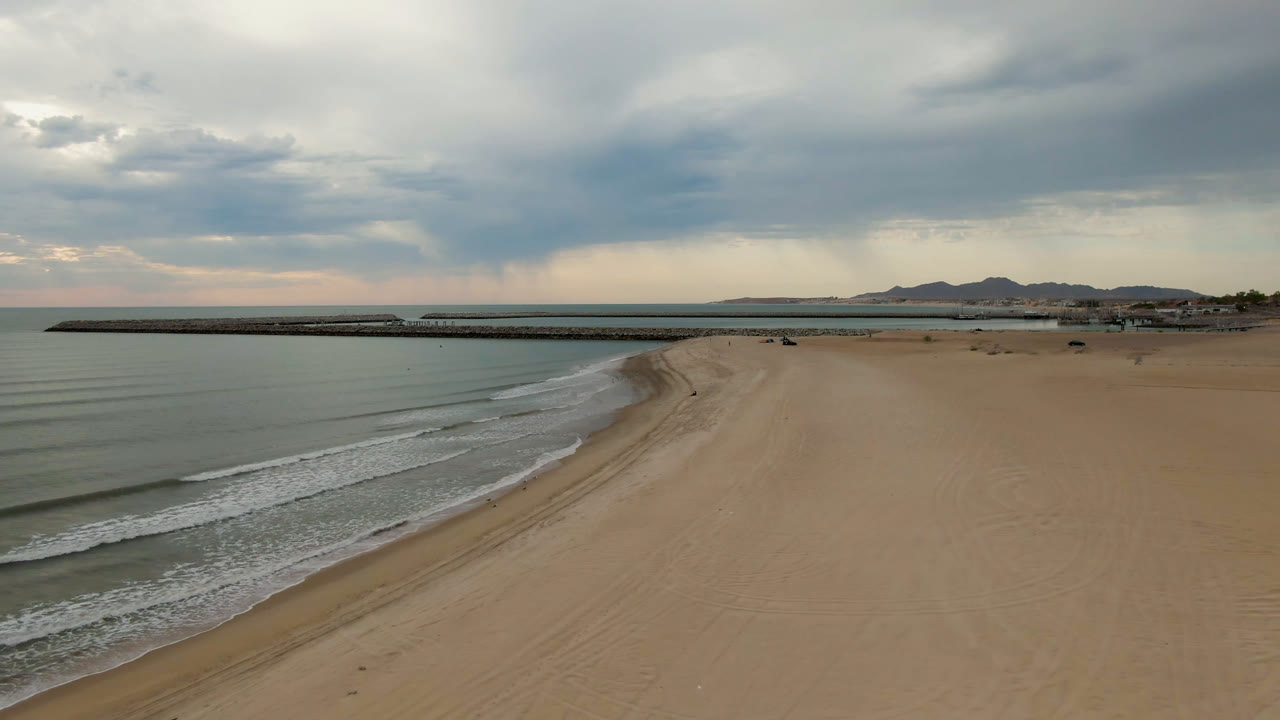 Aerial view in San Felipe, BC, Mexico, showing more defined surf bands along the left edge, wide empty sand on the right, the long pier across the horizon, low hills, layered gray sky, and no visible people.