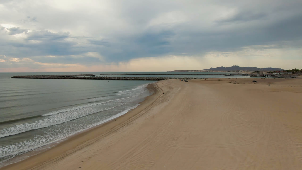 Wide aerial beach frame with surf, open sand, pier, hills, and gray cloud cover.