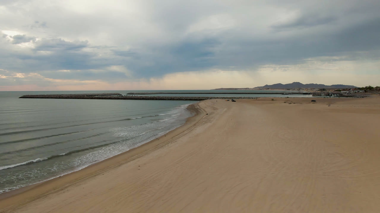 Opening aerial frame over San Felipe, BC, Mexico beach with calm water at left, a long pier across the midground, heavy cloud cover, distant hills, and no visible people.
