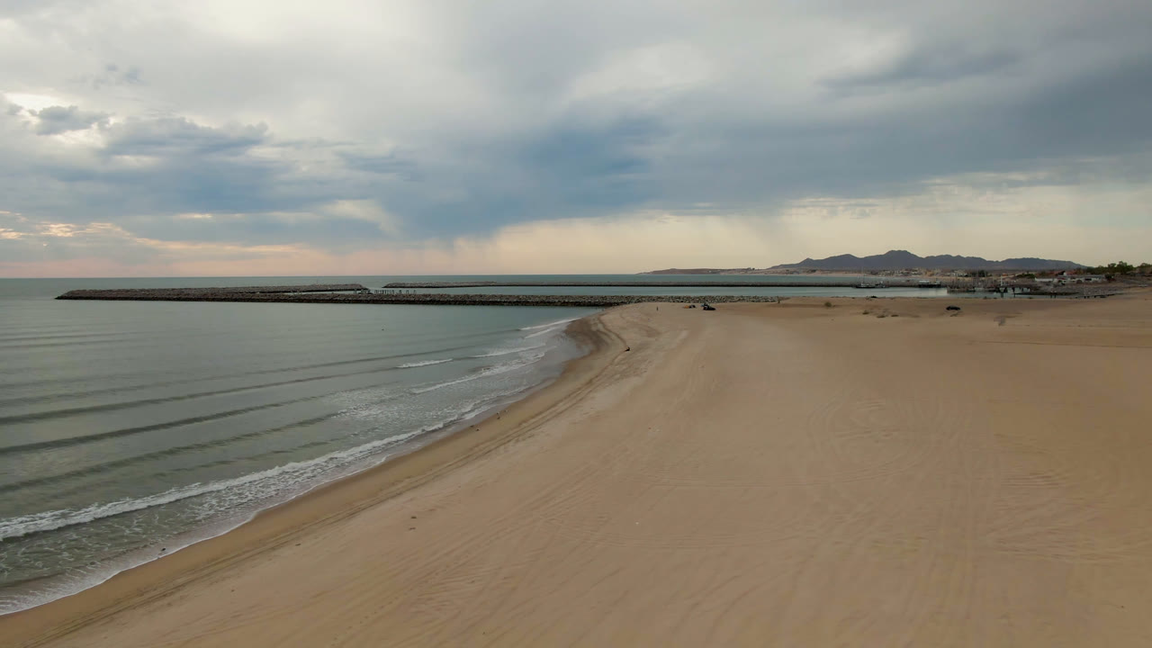Empty beach curving along calm water with pier, clouds, and low mountains.