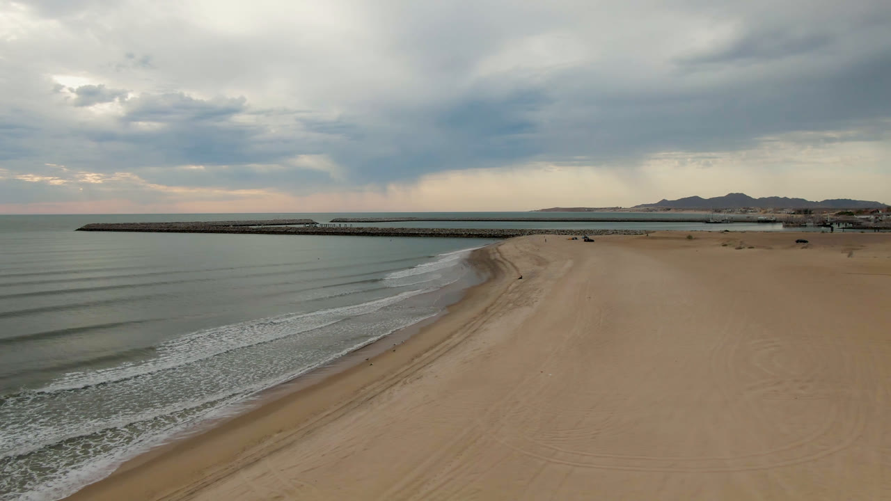 Opening aerial frame over San Felipe, Baja California, Mexico, showing tan beach at right, calm surf at left, a long dark pier across the midground, distant mountains, heavy clouds, and no visible people.