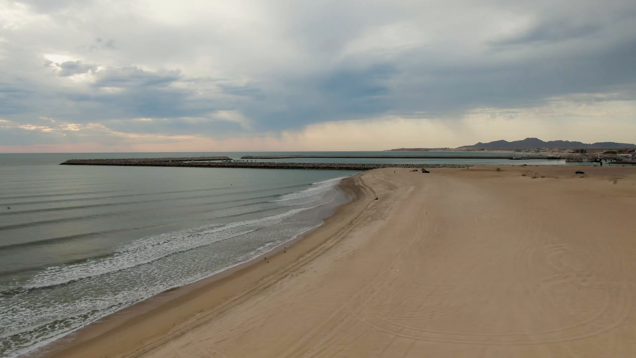 One second into the San Felipe aerial shot, the diagonal shoreline and parallel wave lines are more centered, with the long pier in the midground, low mountains beyond, cloud-filtered light, and no visible people.