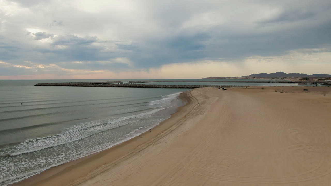 Empty sandy beach beside calm water with a long pier and overcast sky.