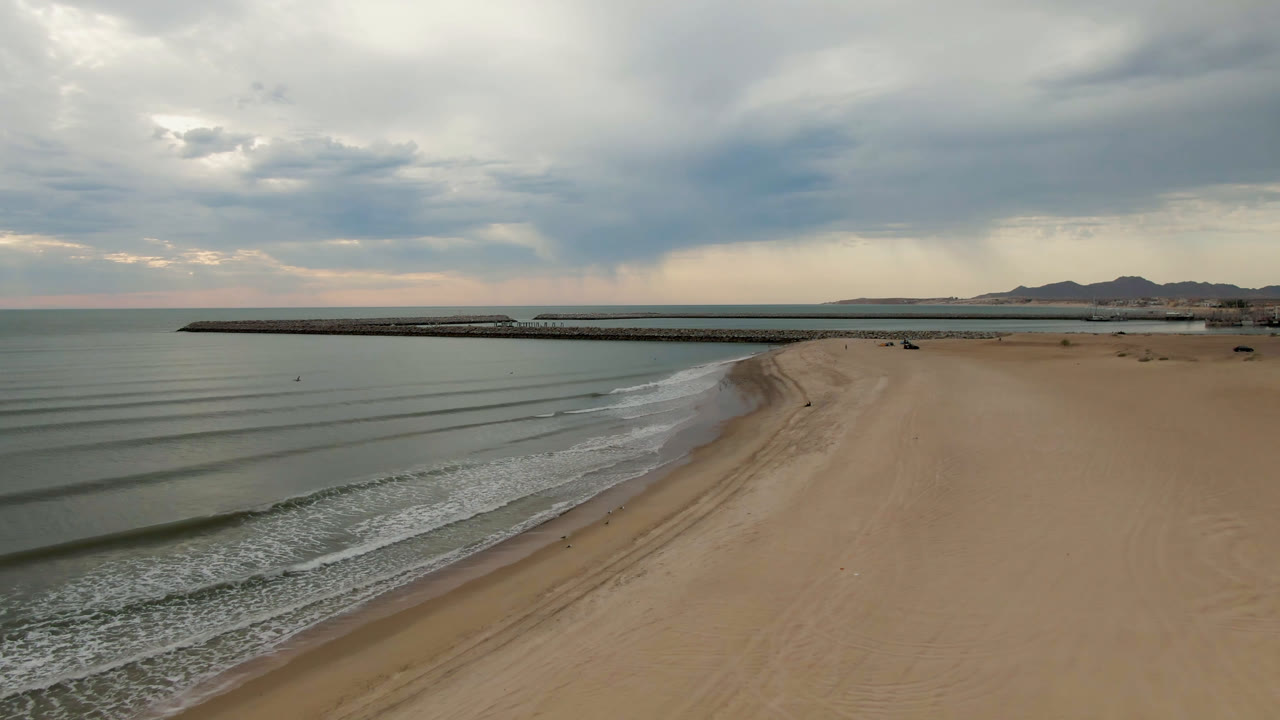 Opening aerial frame over San Felipe beach, with gentle wave lines at left, broad empty sand at right, a long pier across the midground, distant mountains, heavy clouds, and no visible people.