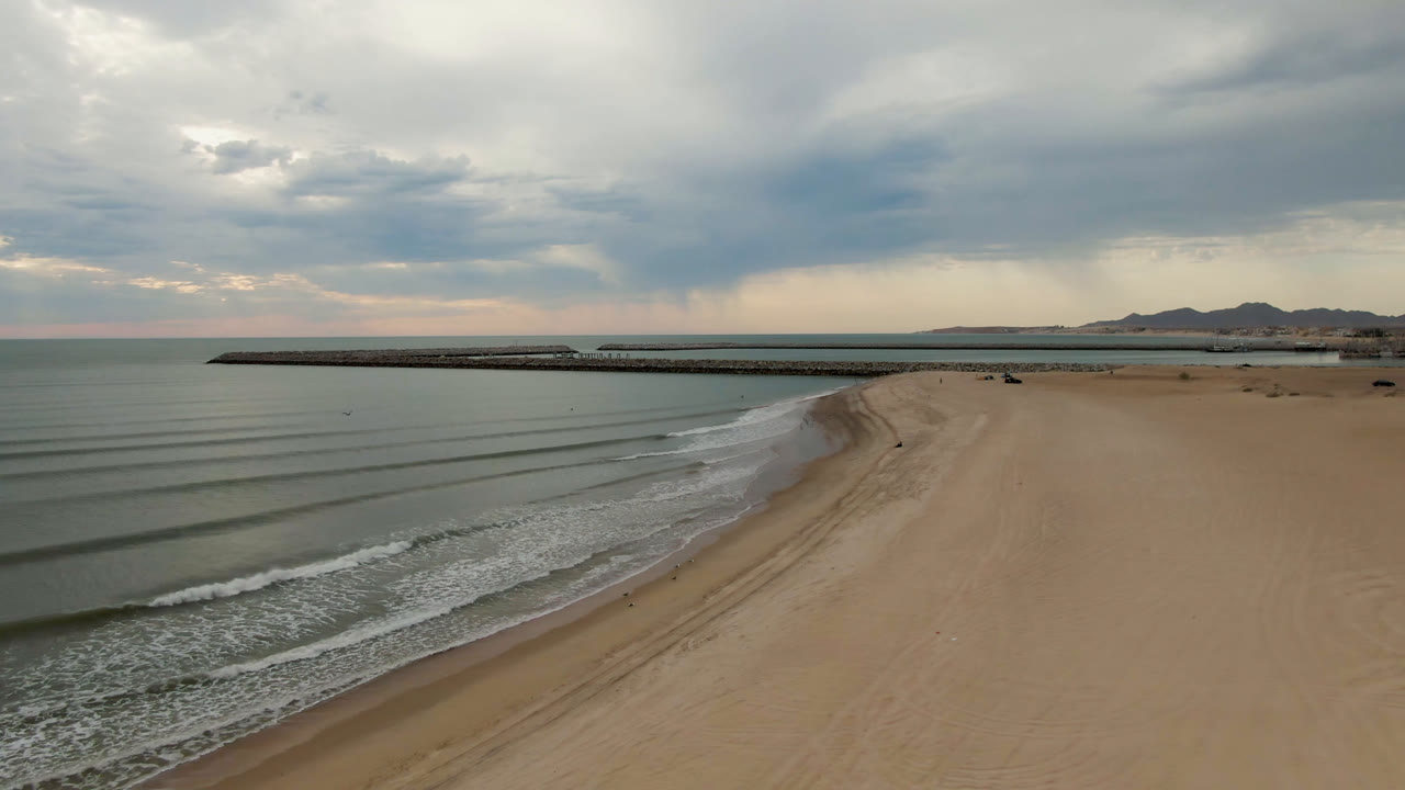 San Felipe shoreline viewed from above at 0:01, with repeating wave bands moving toward the curved sand, the pier stretching across the frame, muted gray-green water, distant mountains, and no visible people.
