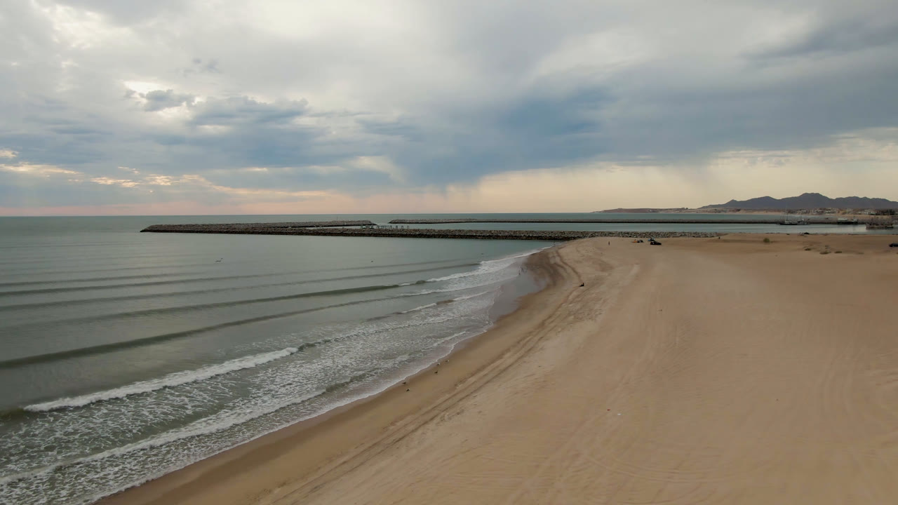 Elevated coastal view with foamy surf, open sand, long pier, and overcast sky.