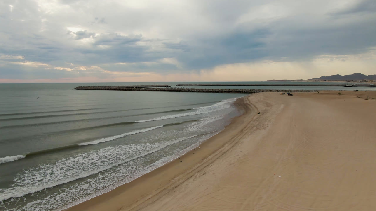 Opening aerial frame over San Felipe, B.C., Mexico, with a broad sandy beach on the right, calm water on the left, a long pier across the background, and no visible people.