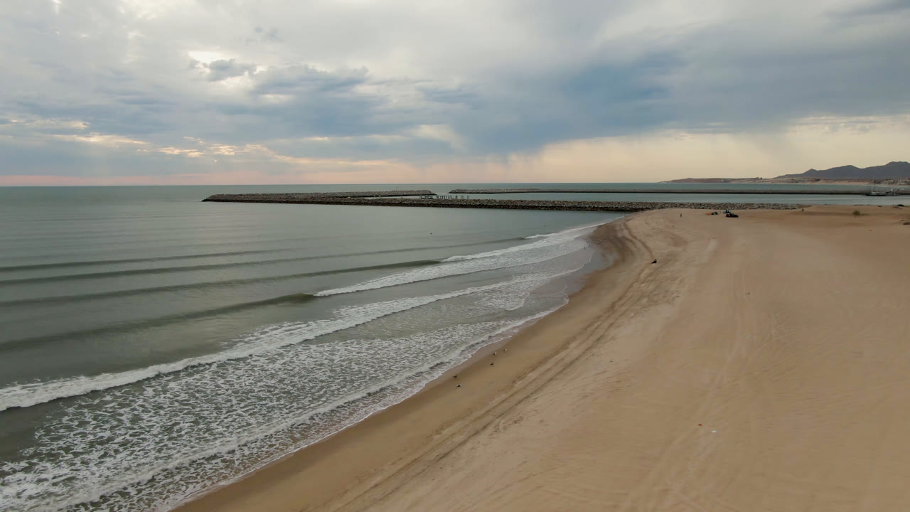 Elevated beach view with surf foam along the shore, open sand, pier, mountains, and layered clouds.
