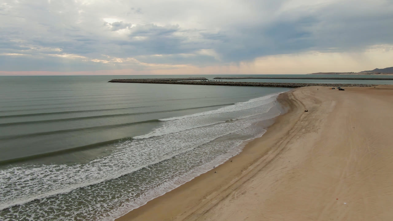Opening aerial frame over San Felipe beach, with green-gray water and parallel surf lines on the left, wide sand on the right, a long pier on the horizon, and no visible people.