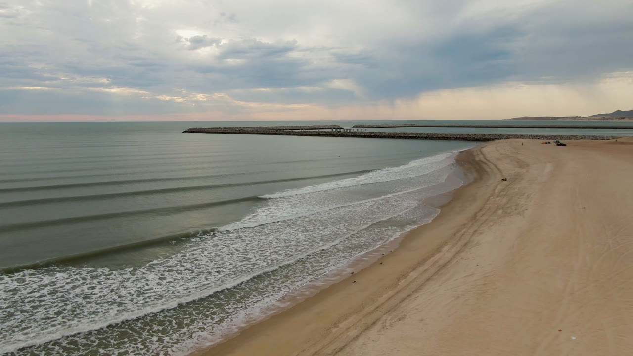 San Felipe aerial beach view one second in, showing wave bands closer to the shoreline, a broad empty sand area, the pier stretching across the upper frame, and no visible people.