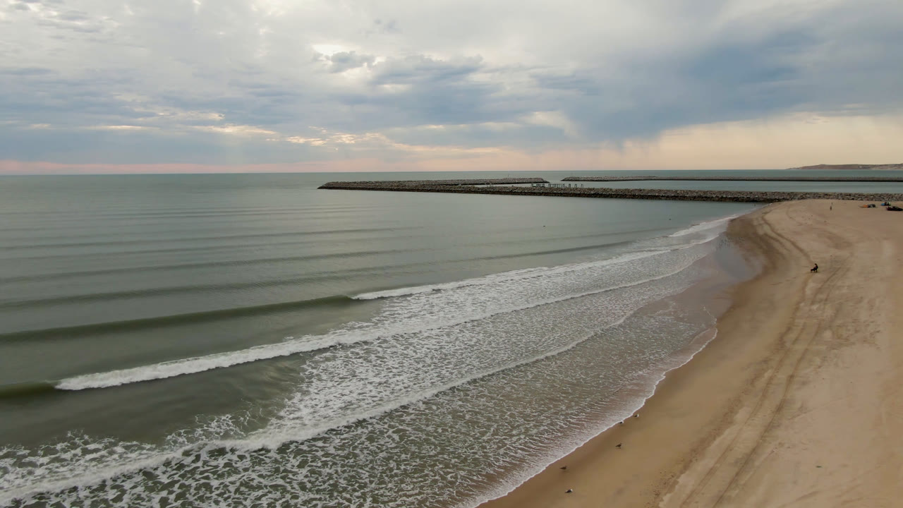 Aerial frame from San Felipe, B.C., Mexico showing parallel waves rolling toward a sandy beach, a long distant pier or breakwater, layered cloud cover, and no visible people.