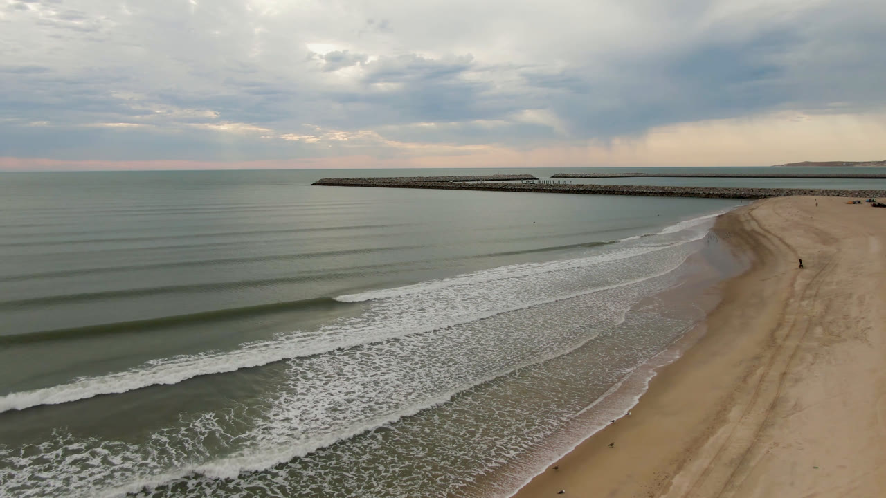 Aerial view in San Felipe, B.C., Mexico with white surf spreading farther up the sand, open green-gray water to the left, a distant pier, low clouds, and no visible people.