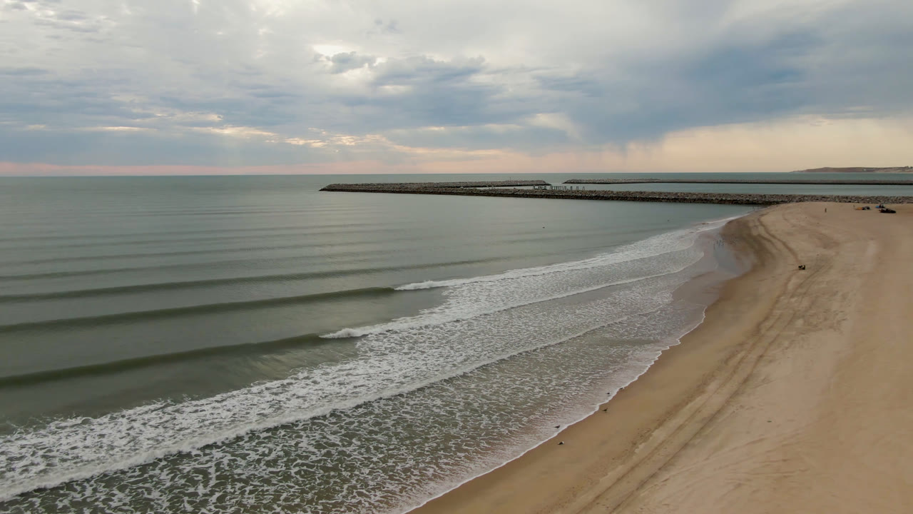 Aerial frame over San Felipe, Mexico, showing parallel surf lines approaching a wide sandy beach, a long pier across the horizon, layered clouds, and no visible people.