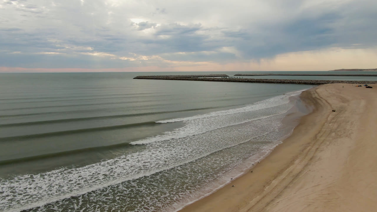 Drone view in San Felipe, Mexico, with green-gray water filling the left side, white surf bands crossing the beach, the pier stretching across the background, and no visible people.
