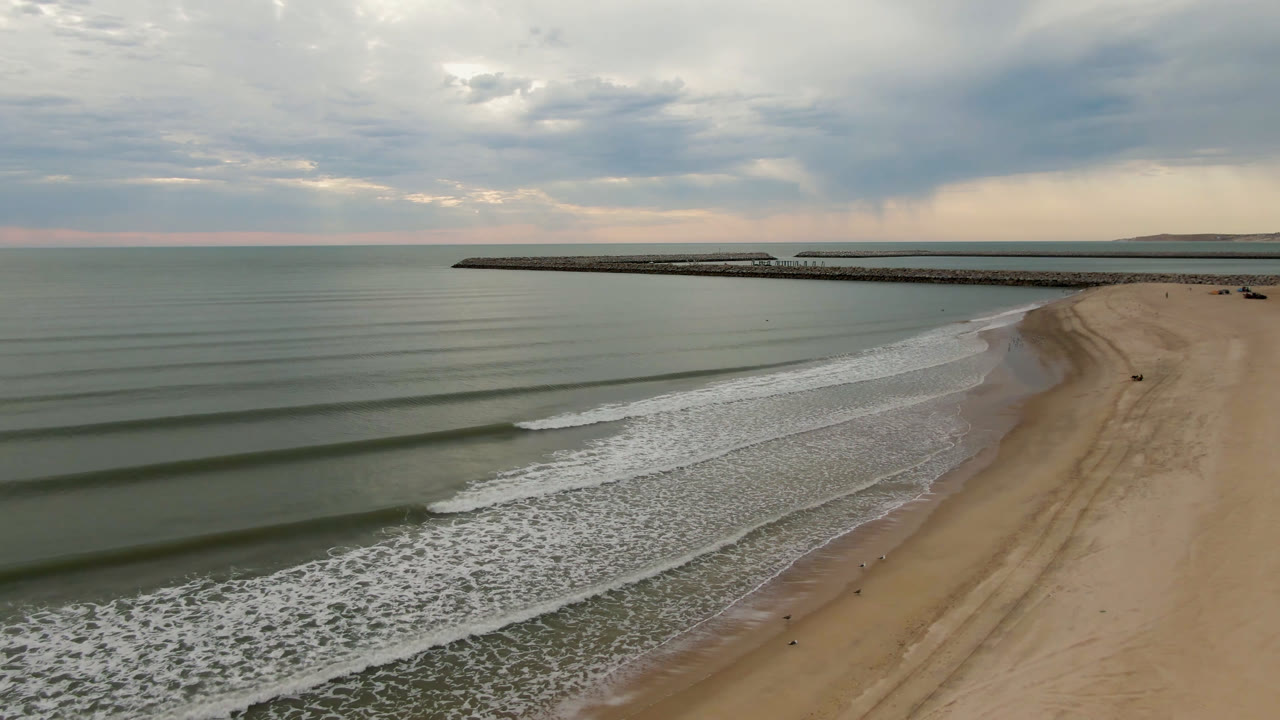 Wide beach aerial with rolling wave bands and a long pier beneath a cloudy sky.
