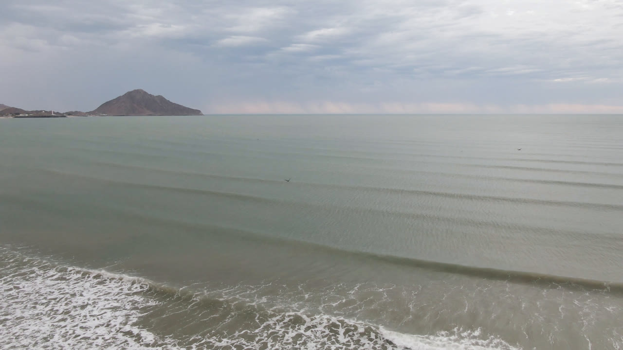 Aerial view over San Felipe, Mexico, with pale ocean, thin wave lines, foamy surf at the bottom edge, distant land on the horizon, and no visible people.