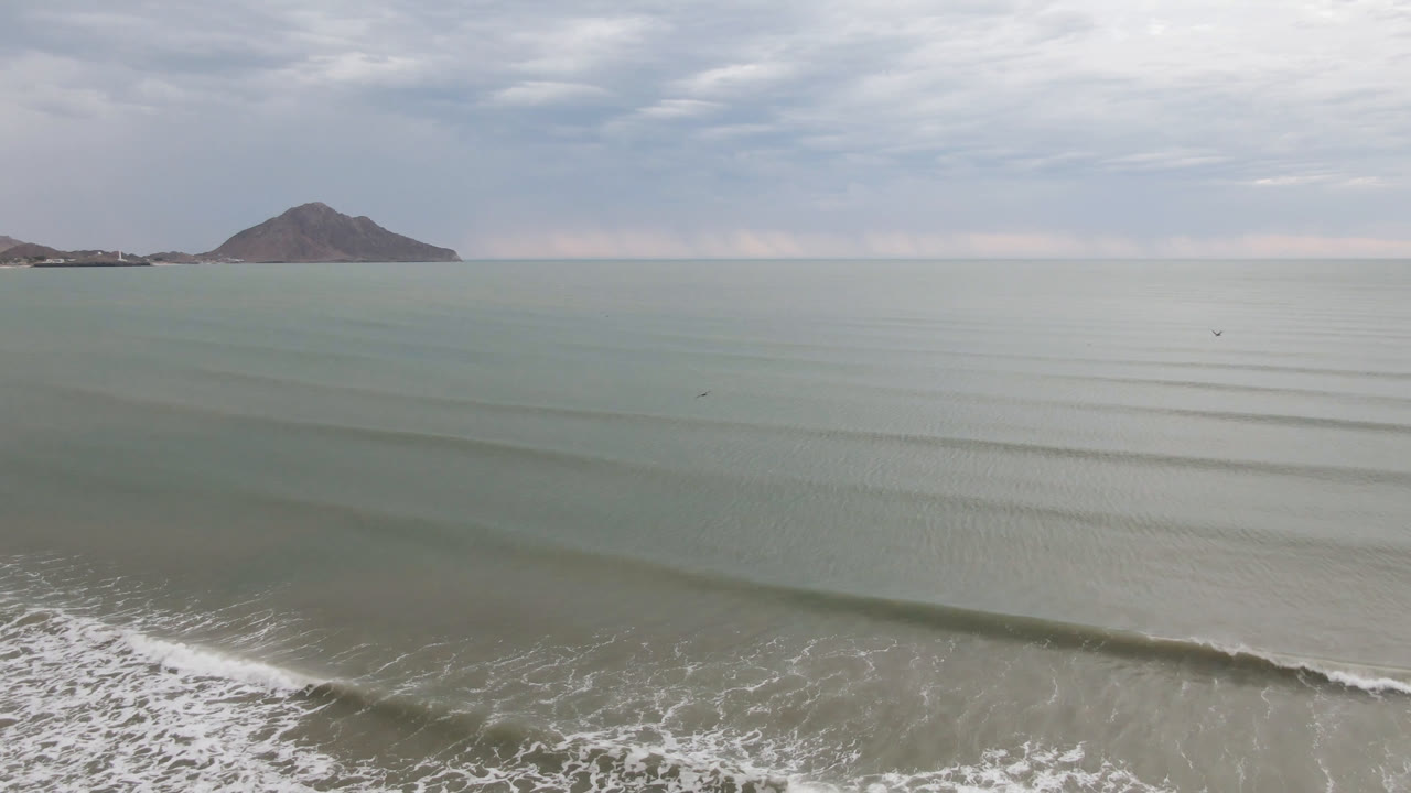 Aerial San Felipe shoreline frame with gentle bands of surf, white foam spreading across the lower foreground, cloudy sky, distant land, and no visible people.