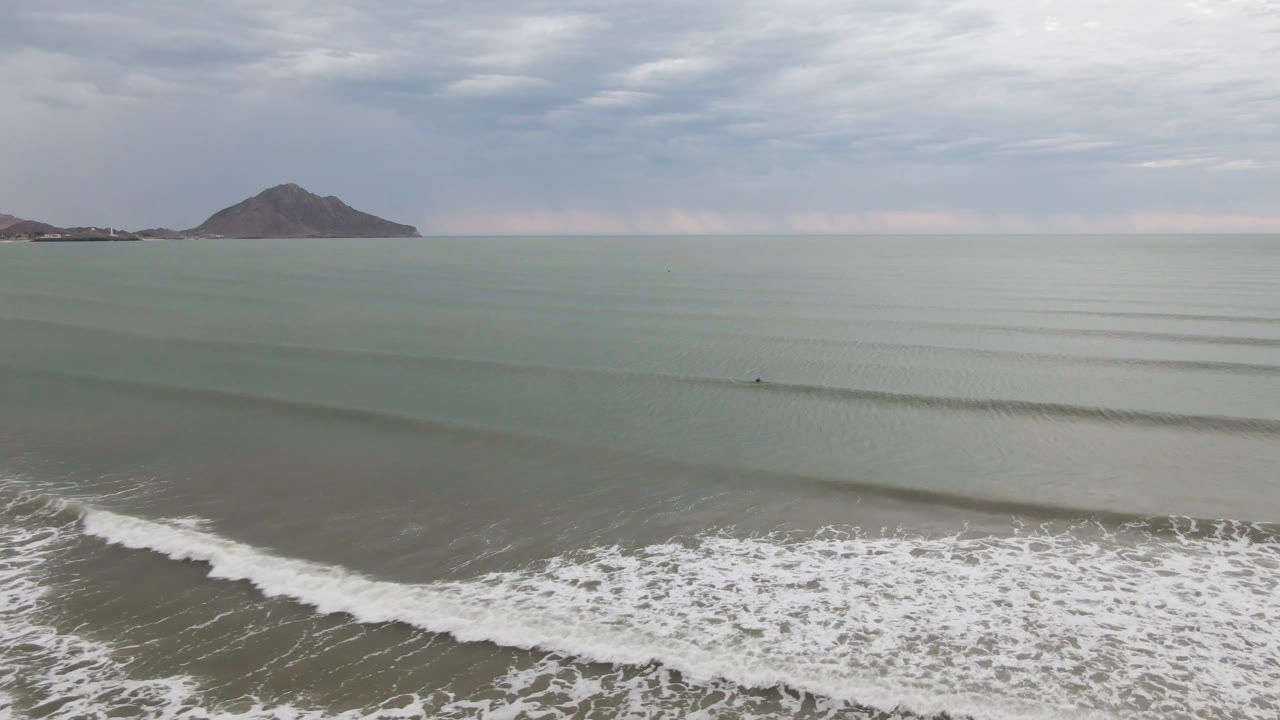 Foamy surf crosses the foreground below calm water, distant shoreline, and a cloudy horizon.