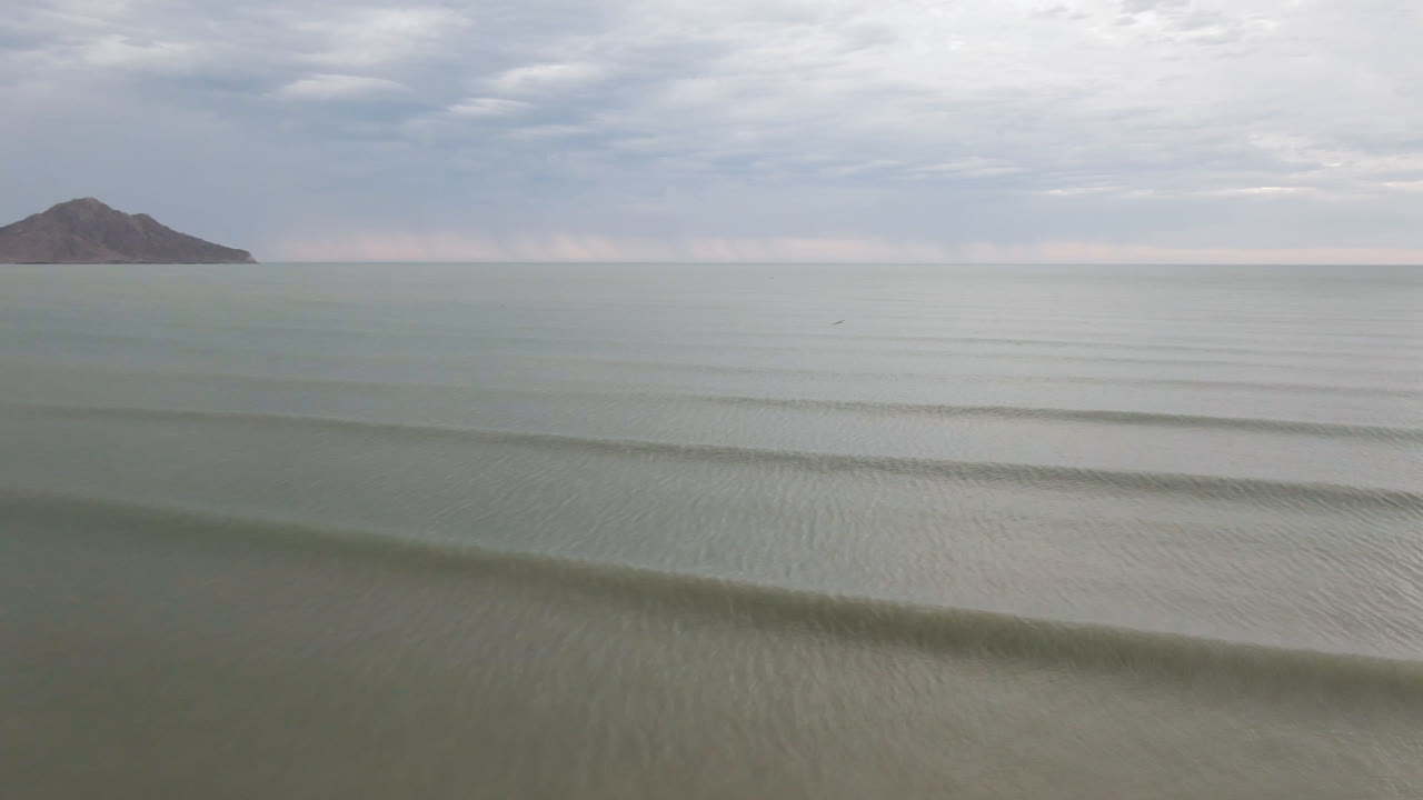 Low waves on a muted ocean with a distant hill under clouds.