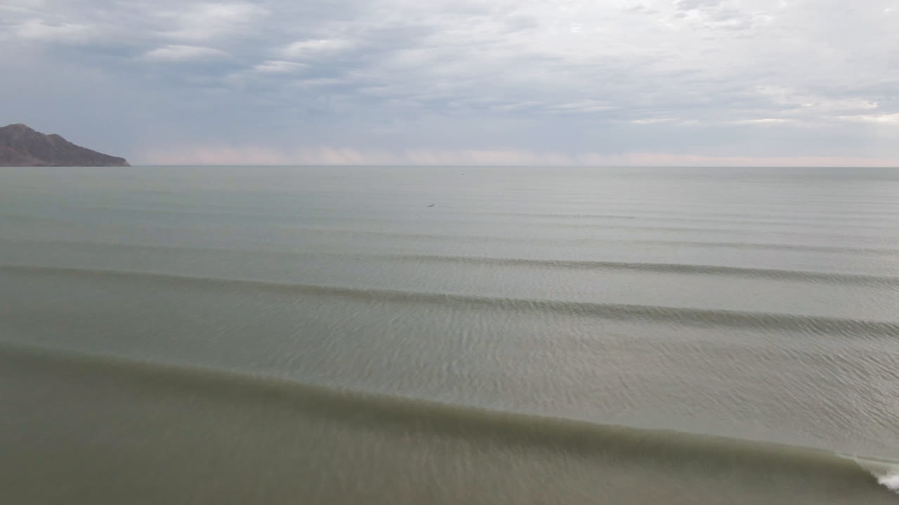 San Felipe coastal water stretches toward a cloudy horizon, with parallel ripple lines in the foreground, the offshore hill closer to center, and no visible people.
