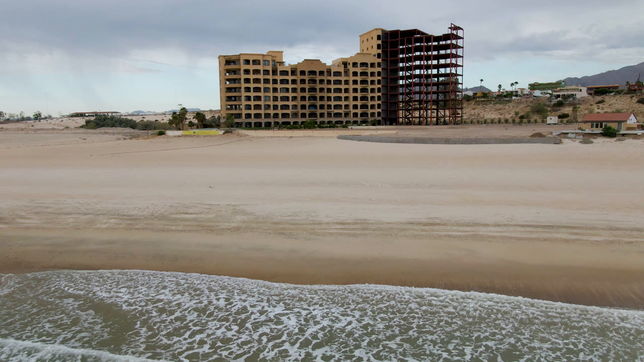 Wide beach with surf in front of a large tan building and exposed framing.