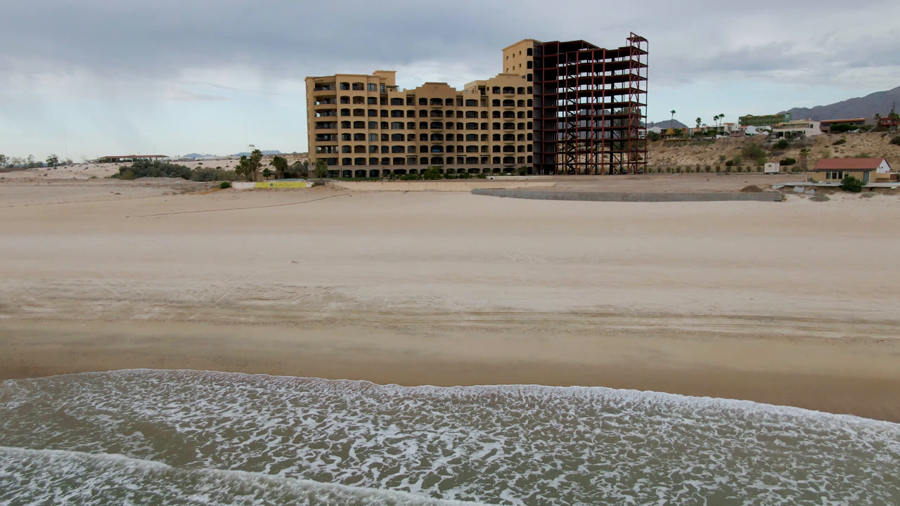 Second frame in San Felipe, Baja California, Mexico with the surf line lower in frame, broad empty sand across the center, the large tan building and exposed frame above it, and no visible people.