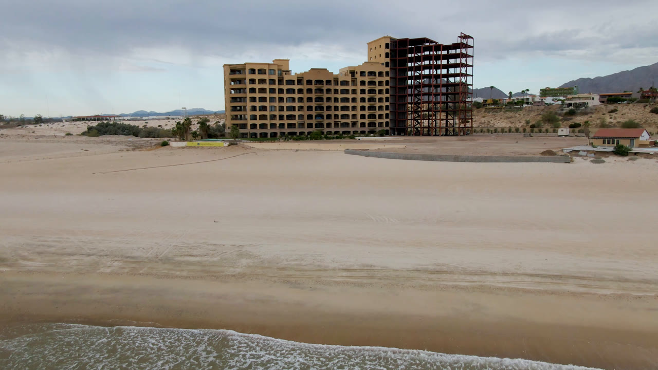 Wide beach with waves, sand, unfinished beachfront building, and cloudy sky.