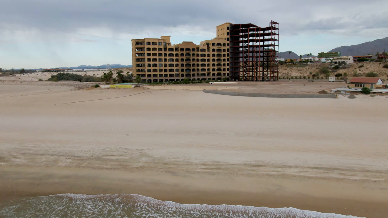 A broad San Felipe shoreline remains empty, with pale sand across the foreground, a large unfinished building near the center, hills behind it, and no visible people.