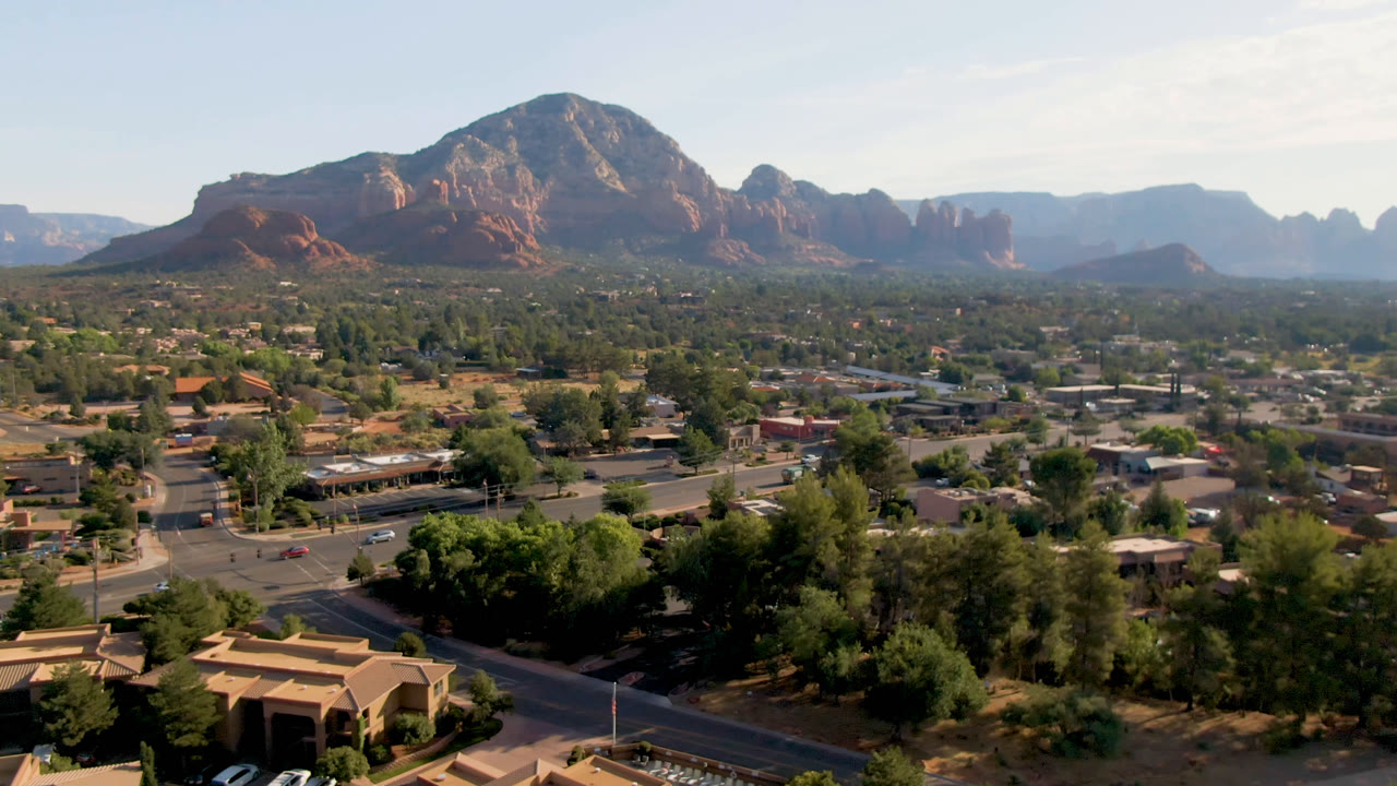 Sedona aerial frame with foreground roads and buildings, dense green tree cover, and a large red rock formation rising behind the town with no visible people.