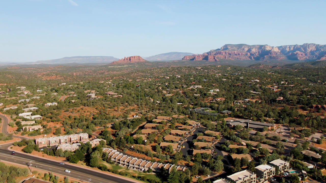 Wide 0:00 aerial frame over Sedona, Arizona with a road and low buildings in the foreground, tree-filled town in the midground, red rock mountains beyond, and no visible people.