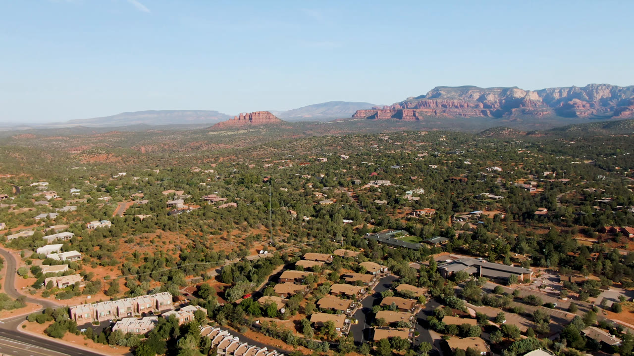 Wide aerial view over Sedona, Arizona, with low buildings, winding roads, green tree cover, red soil, distant red rock formations, and no visible people.