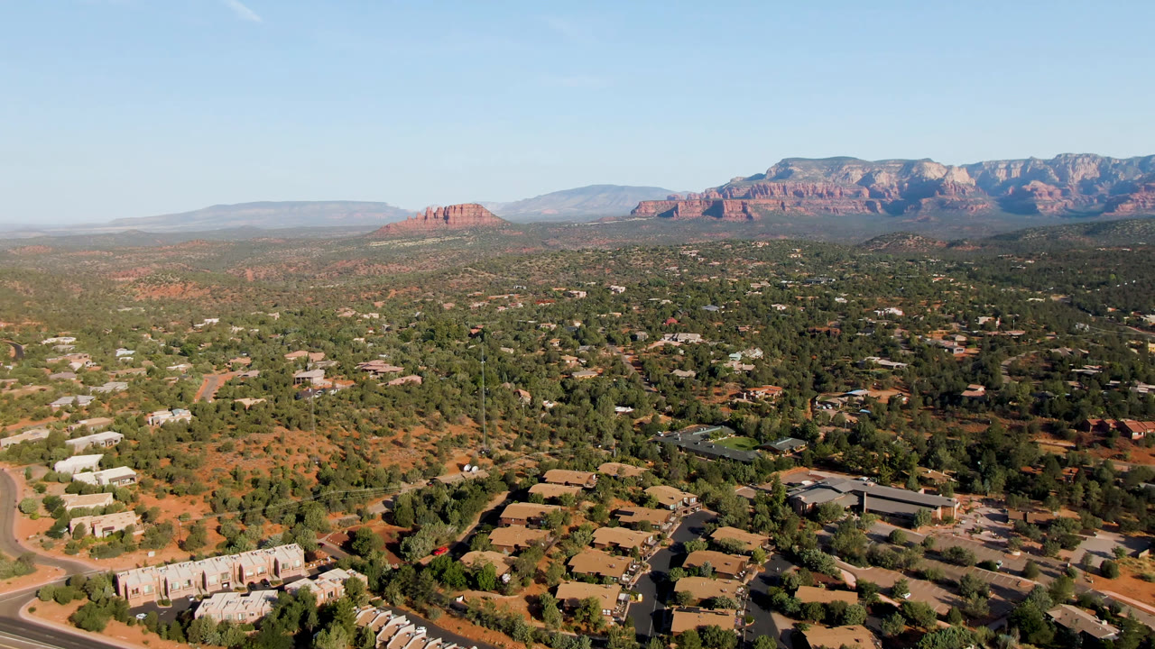 Aerial frame over Sedona, Arizona, showing residential and low-rise structures among trees, pale roads, red desert ground, distant mesas, and no visible people.