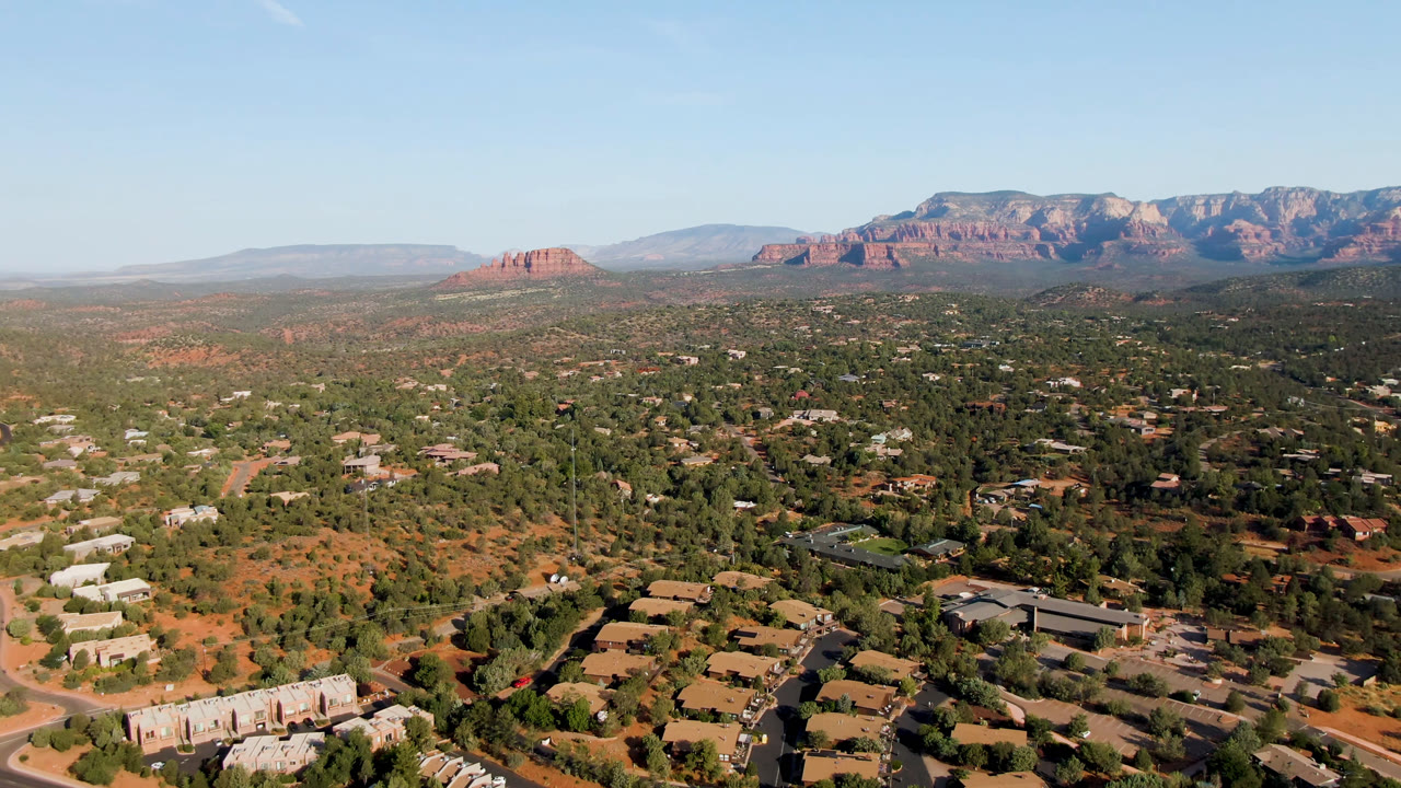High aerial view of Sedona, Arizona, with town blocks in the foreground, scattered tree cover through the middle distance, red rock formations along the horizon, and no visible people.