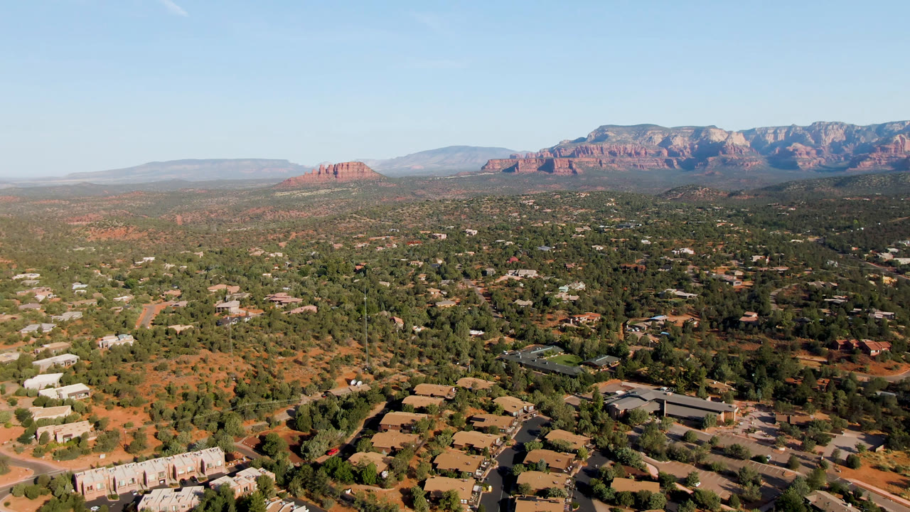 Wide drone view of Sedona buildings, curved roads, trees, and distant red mountains.