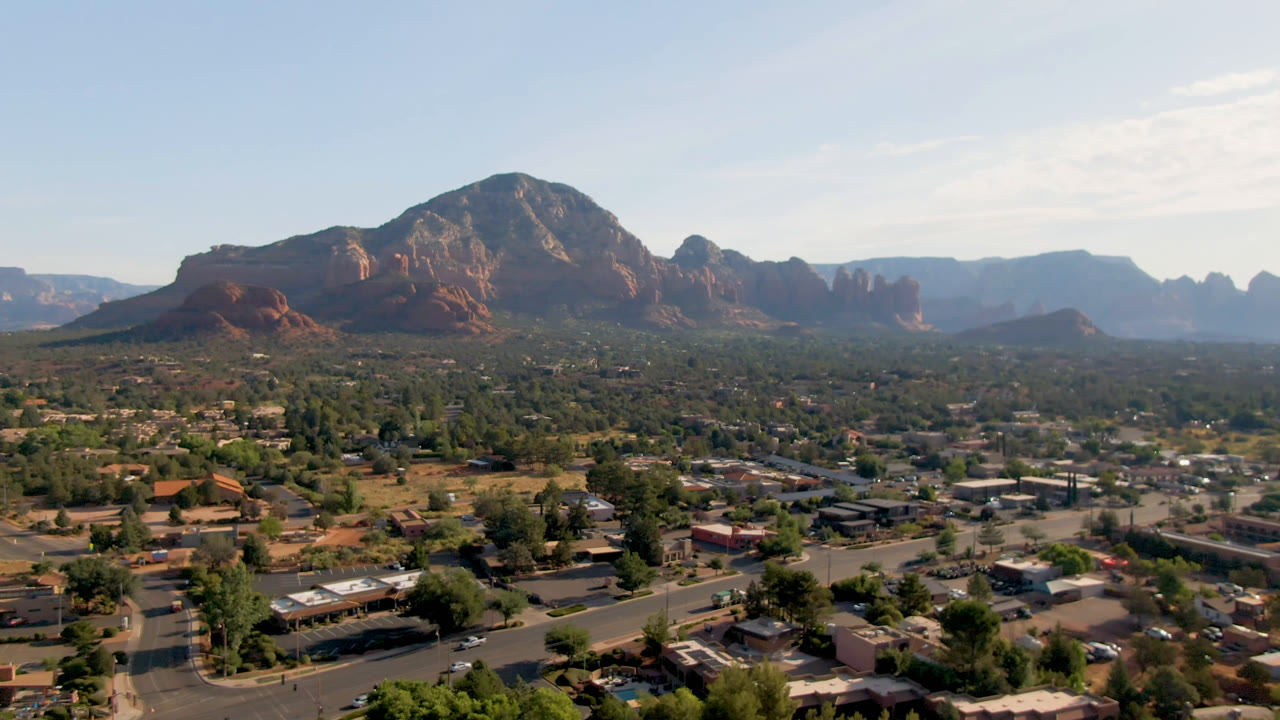 Opening frame of Sedona, Arizona from above, with roads, low buildings, scattered trees, red rock mountains, and no visible people.