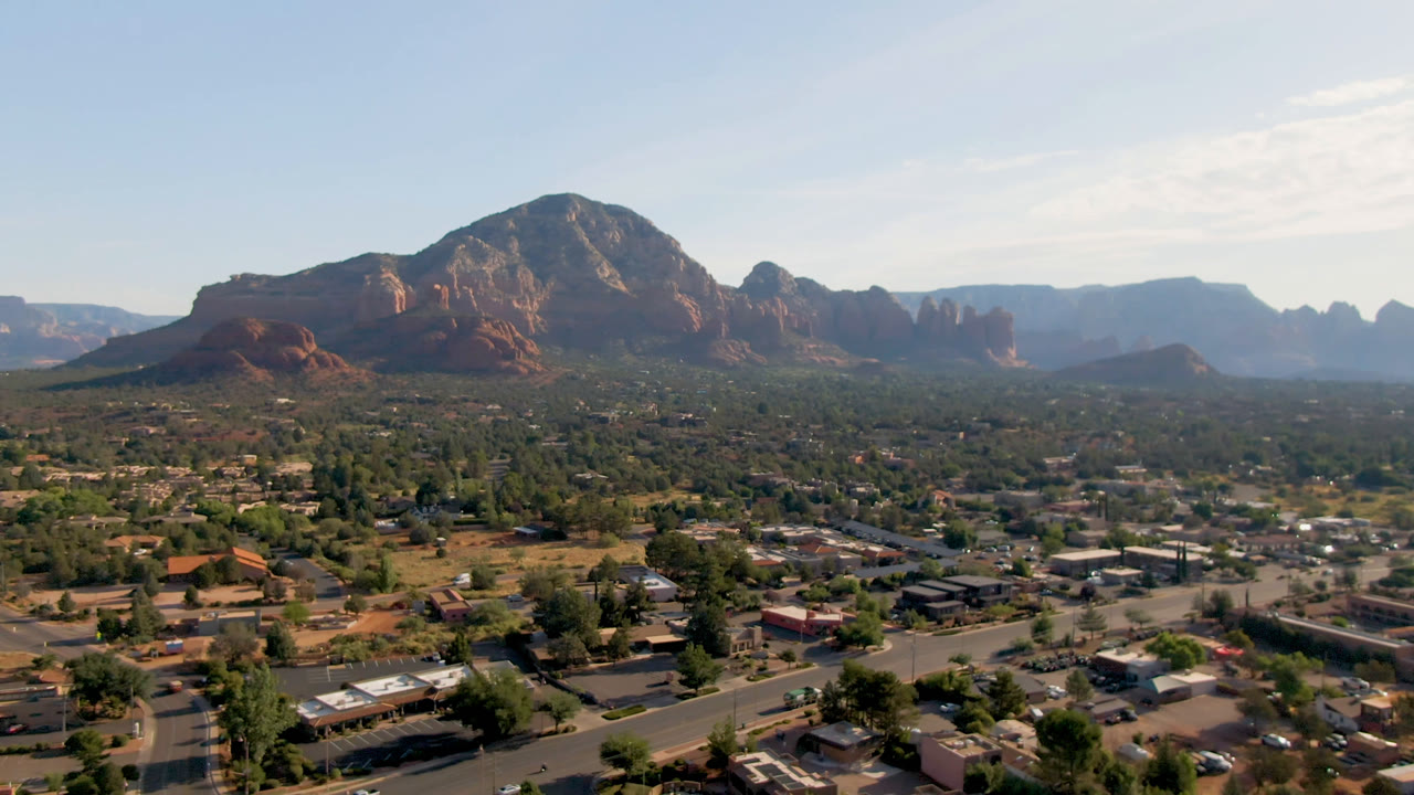 Sedona, Arizona view showing a slightly later wide aerial angle over roadways, roofs, green tree cover, layered red rocks, and no visible people.