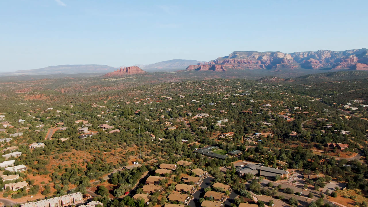 Opening aerial frame over Sedona, Arizona showing scattered homes, green desert vegetation, red earth, and a red rock mountain range beneath a clear blue sky with no visible people.
