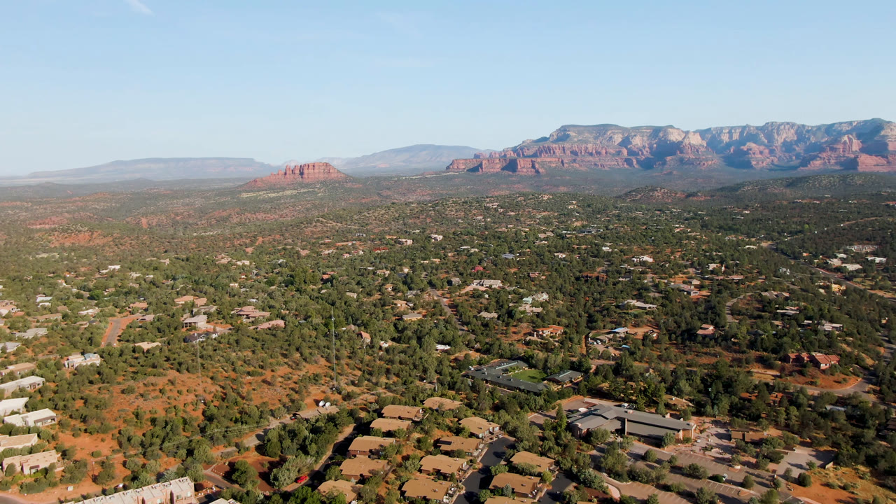 Aerial view across trees and homes toward red rock bluffs and a blue sky.