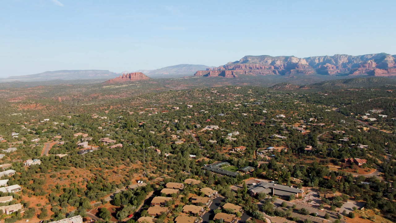 Midwide aerial frame over Sedona, Arizona showing more open red terrain at left, clusters of homes and trees below, and the mountain range stretching across the background with no visible people.