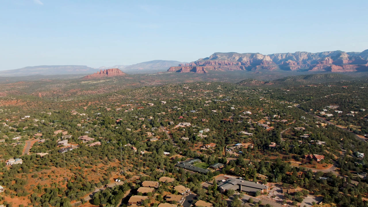 Wide aerial view over Sedona, Arizona, with scattered buildings, green desert vegetation, red rock formations on the horizon, clear blue sky, and no visible people.