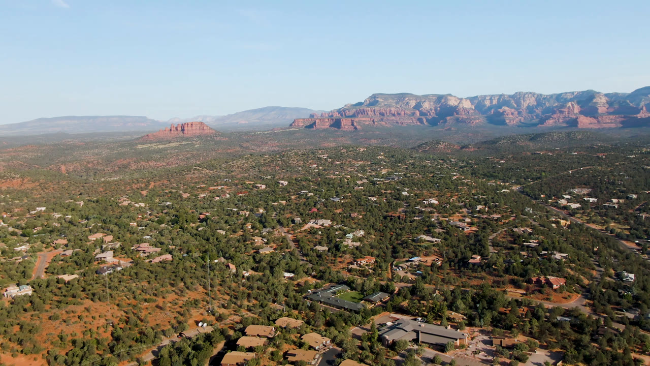 High aerial frame over Sedona showing more of the central town grid, desert trees, low buildings, red rock cliffs across the background, and no visible people.