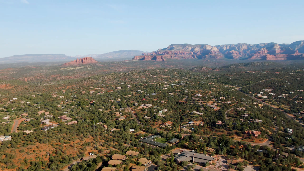Aerial view of Sedona with roads and buildings in the lower frame, patches of red soil and green trees through the midground, distant red rock ridges, and no visible people.